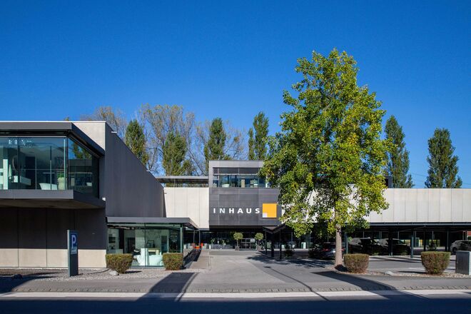 Ein modernes Gebäude mit der Aufschrift "INHAUS". Es hat eine klare, geradlinige Architektur mit großen Fenstern. Ein großer Baum steht auf dem Vorplatz, der von Pflanzen gesäumt ist. Ein modernes Gebäude mit der Aufschrift "INHAUS". Es hat eine klare, geradlinige Architektur mit großen Fenstern. Ein großer Baum steht auf dem Vorplatz, der von Pflanzen gesäumt ist.